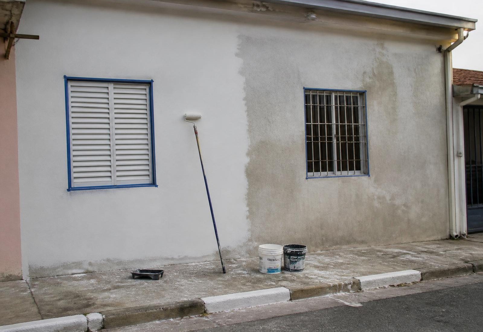 Fachada de casa em pintura, com metade da parede já pintada e latas de tinta na calçada